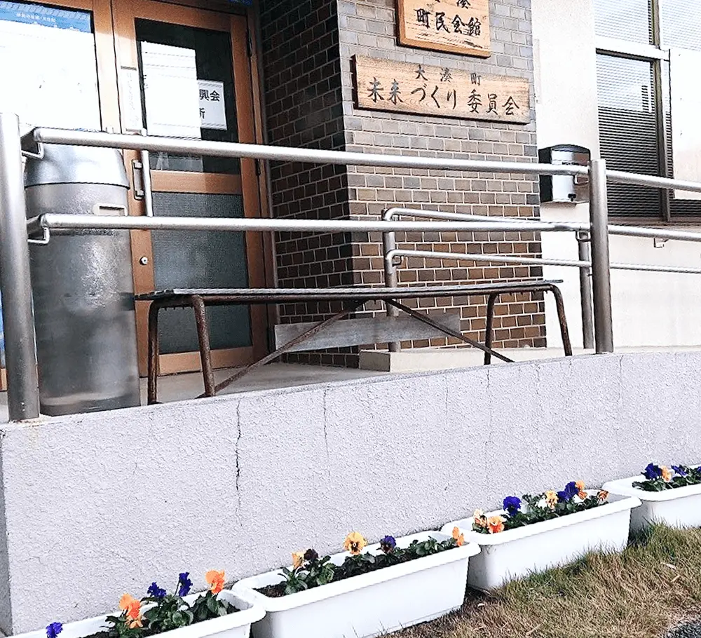 大湊町民会館と未来づくり委員会の木製看板が掲げられた建物入口。バリアフリー対応の手すり付きスロープがあり、手前には紫やオレンジ色のパンジーが植えられた白いプランターが並んでいる